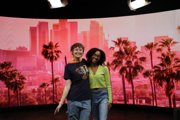 2 hosts standing in front of pink cityscape background.